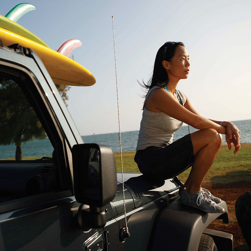 Female student sitting on a jeep before surfing, with ocean in background.